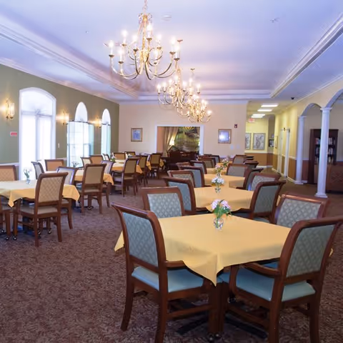 A spacious dining room with multiple tables covered in yellow tablecloths, each adorned with small floral centerpieces. The room features elegant chandeliers hanging from the ceiling, large windows with arched tops letting in natural light, and a carpeted floor. The walls are painted in soft pastel colors, and there are framed pictures and decorative elements on the walls.