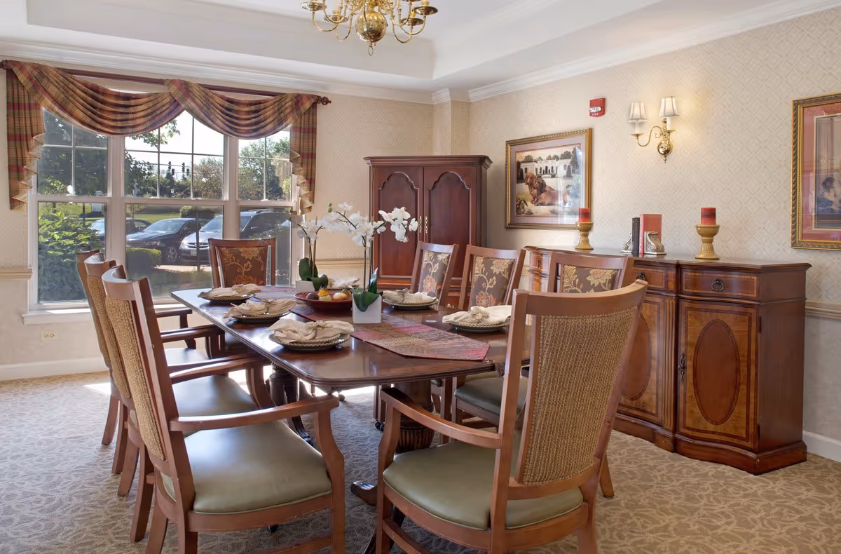 Sunlit formal dining room with a wooden table set with place settings and an orchid centerpiece, upholstered chairs, a sideboard, and a large window with a draped valance.