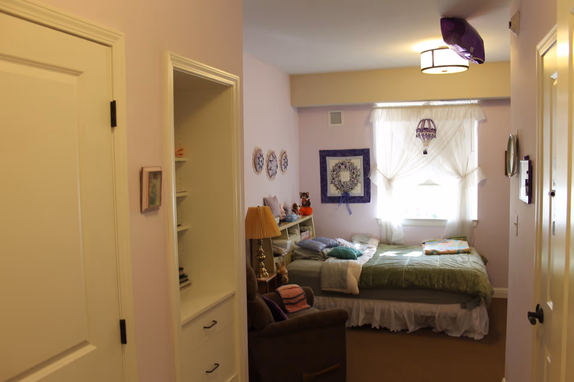 A cozy bedroom with a single bed covered in green bedding and pillows, a window with sheer white curtains letting in natural light, a brown armchair with a folded blanket, a small lamp on a side table, and decorative wall hangings including a framed quilt and circular art pieces.