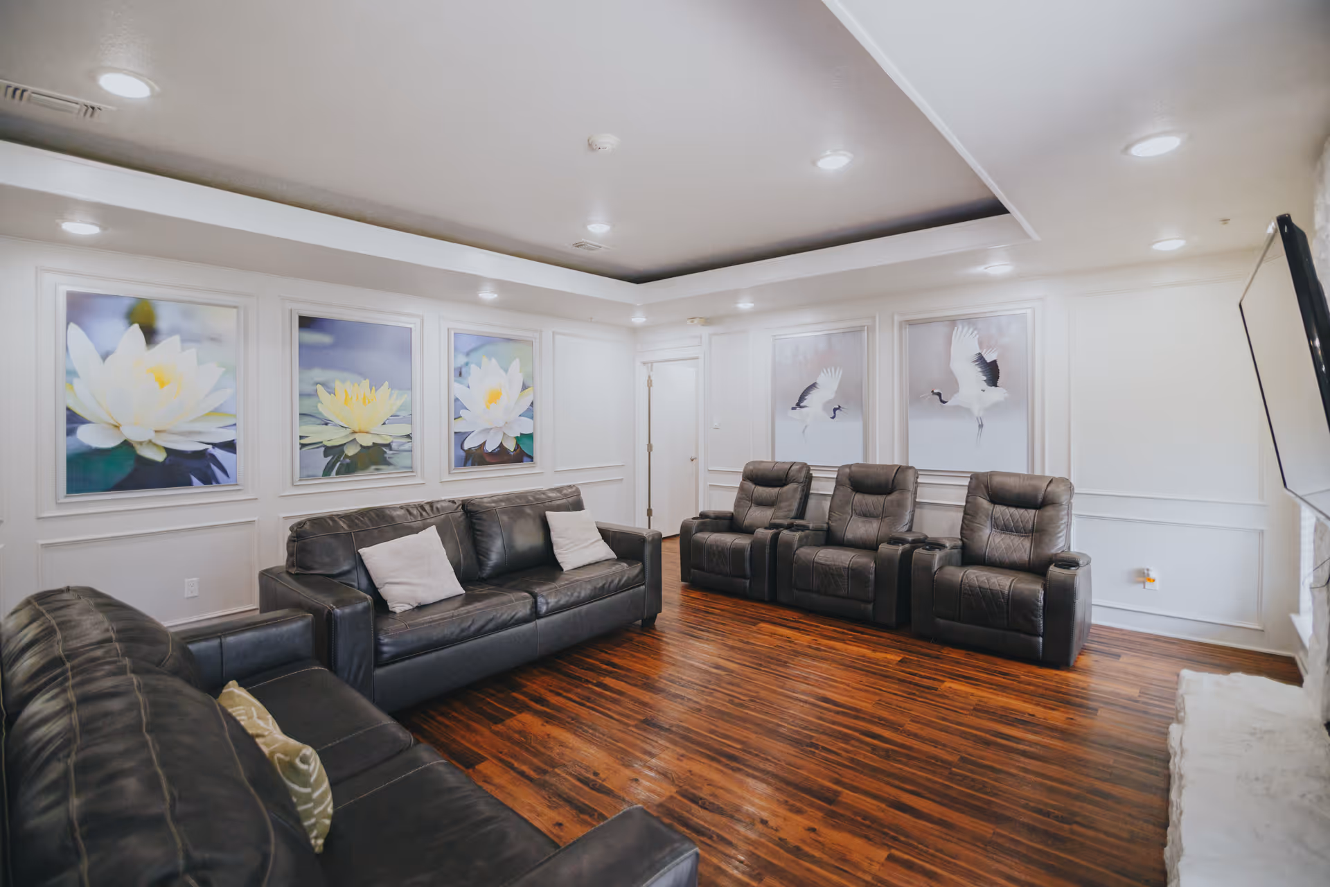 A cozy living room with dark leather sofas and recliner chairs arranged on a polished wooden floor. The walls are decorated with framed pictures of white flowers and birds. The ceiling has recessed lighting, and a flat-screen TV is mounted on the right wall.