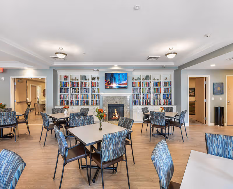 Bright common dining room with tables and patterned chairs, a fireplace flanked by bookshelves and a wall-mounted TV.