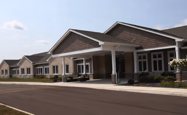 Exterior view of a single-story assisted living facility building with a covered entrance, multiple windows, and a paved driveway under a partly cloudy sky.