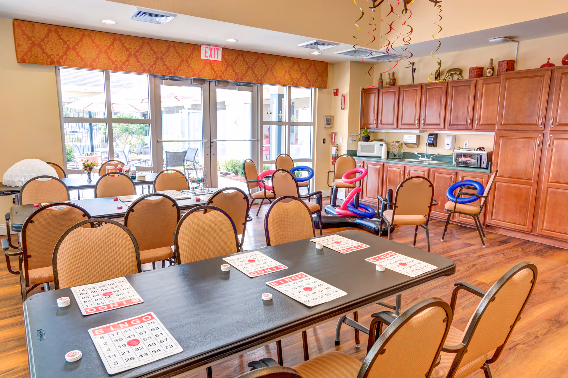 Community activity room with tables and chairs set up for bingo, a kitchenette with wooden cabinets along the back wall, and glass doors leading outside.