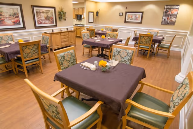 A dining room with several wooden tables covered with dark tablecloths, each set with napkins, utensils, and small flower centerpieces. The room has wooden flooring, floral upholstered chairs, framed artwork on the walls, and soft lighting.