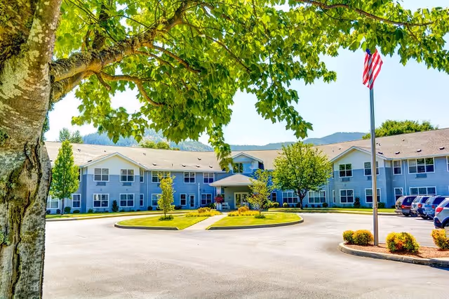 Front exterior view of a two-story assisted living facility building with a circular driveway, landscaped greenery, and an American flag on a flagpole. A large tree with green leaves is in the foreground.