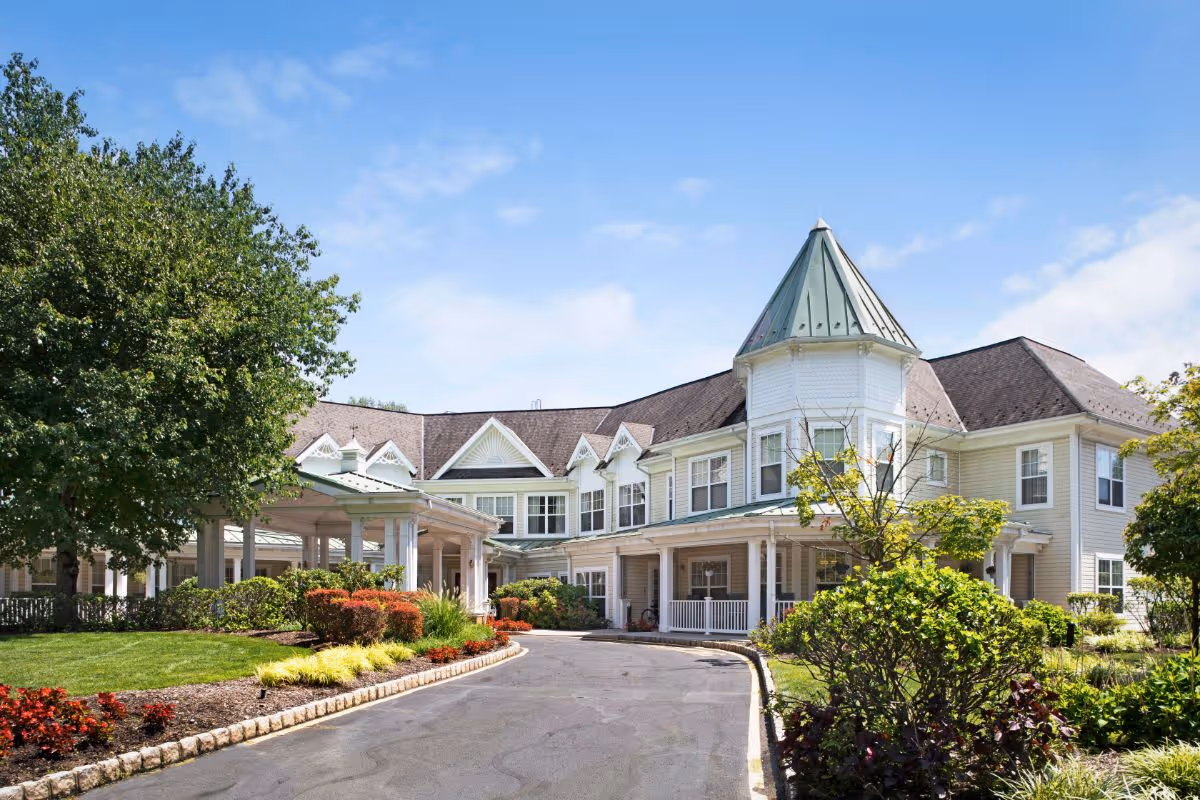 Exterior view of a senior living facility building with a turret, covered entrance, and well-maintained landscaping including trees, bushes, and flowers under a blue sky.
