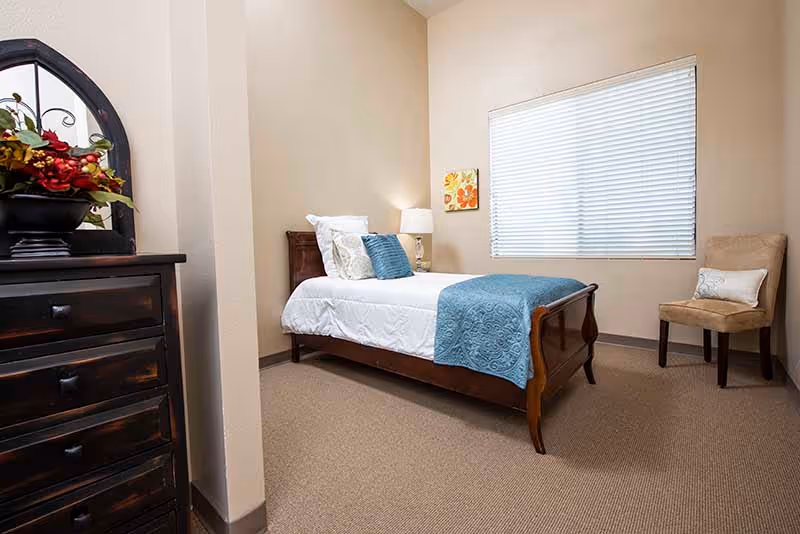 A single bed with white linens and a blue throw in a tidy furnished bedroom with a dresser, nightstand, chair, and window blinds.