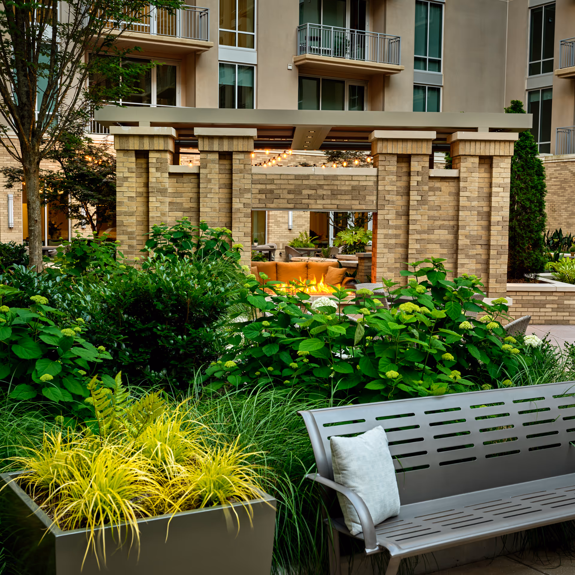 Outdoor seating area in a garden courtyard of a senior living facility with lush green plants, a modern metal bench with a cushion, and a brick structure with a fire pit and string lights in the background. The building with balconies is visible behind the courtyard.