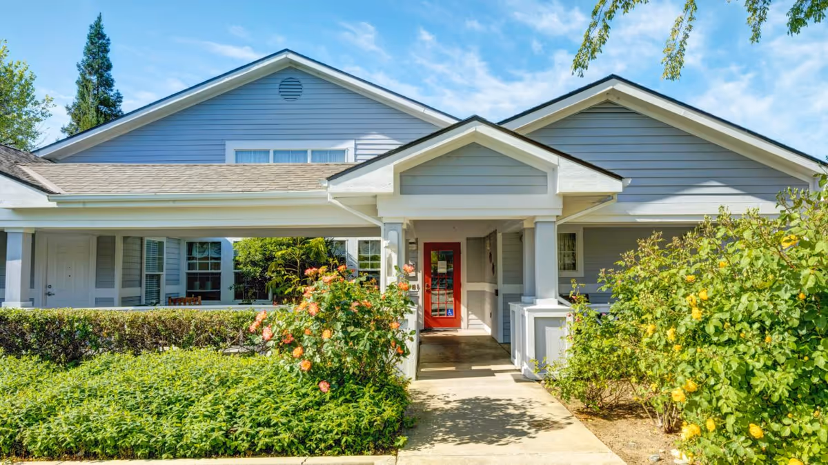Front exterior view of a single-story building with gray siding and a red door entrance, surrounded by green bushes and flowering plants under a blue sky.