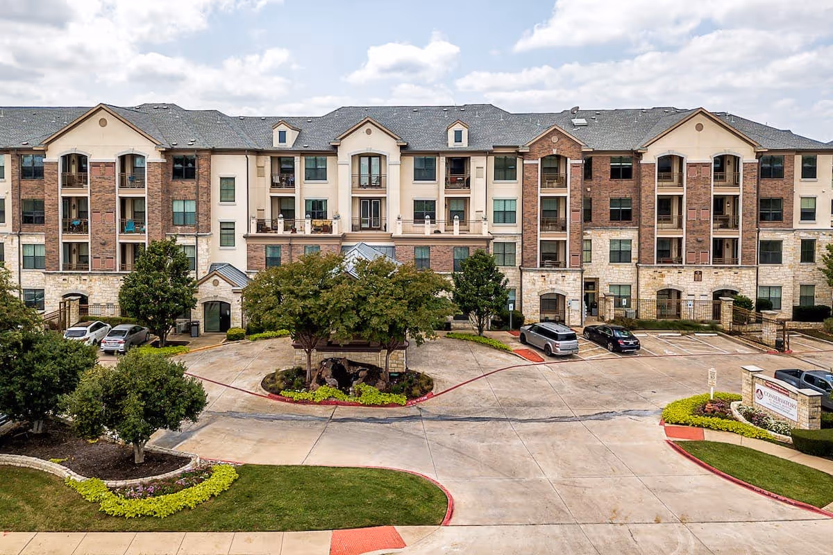 Front exterior view of a multi-story senior living facility named Conservatory At North Austin, featuring a large parking area, landscaped greenery, and multiple balconies on the building.