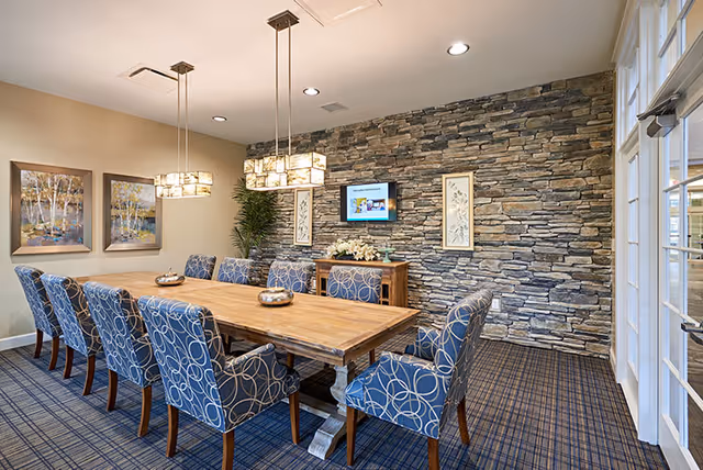 A dining room with a long wooden table surrounded by ten blue upholstered chairs with white circular patterns. The room features a stone accent wall with a mounted flat-screen TV and two framed artworks. Two modern pendant lights hang above the table, and there are two landscape paintings on the adjacent wall. The floor is carpeted with a blue and brown checkered pattern, and there is a glass door on the right side.