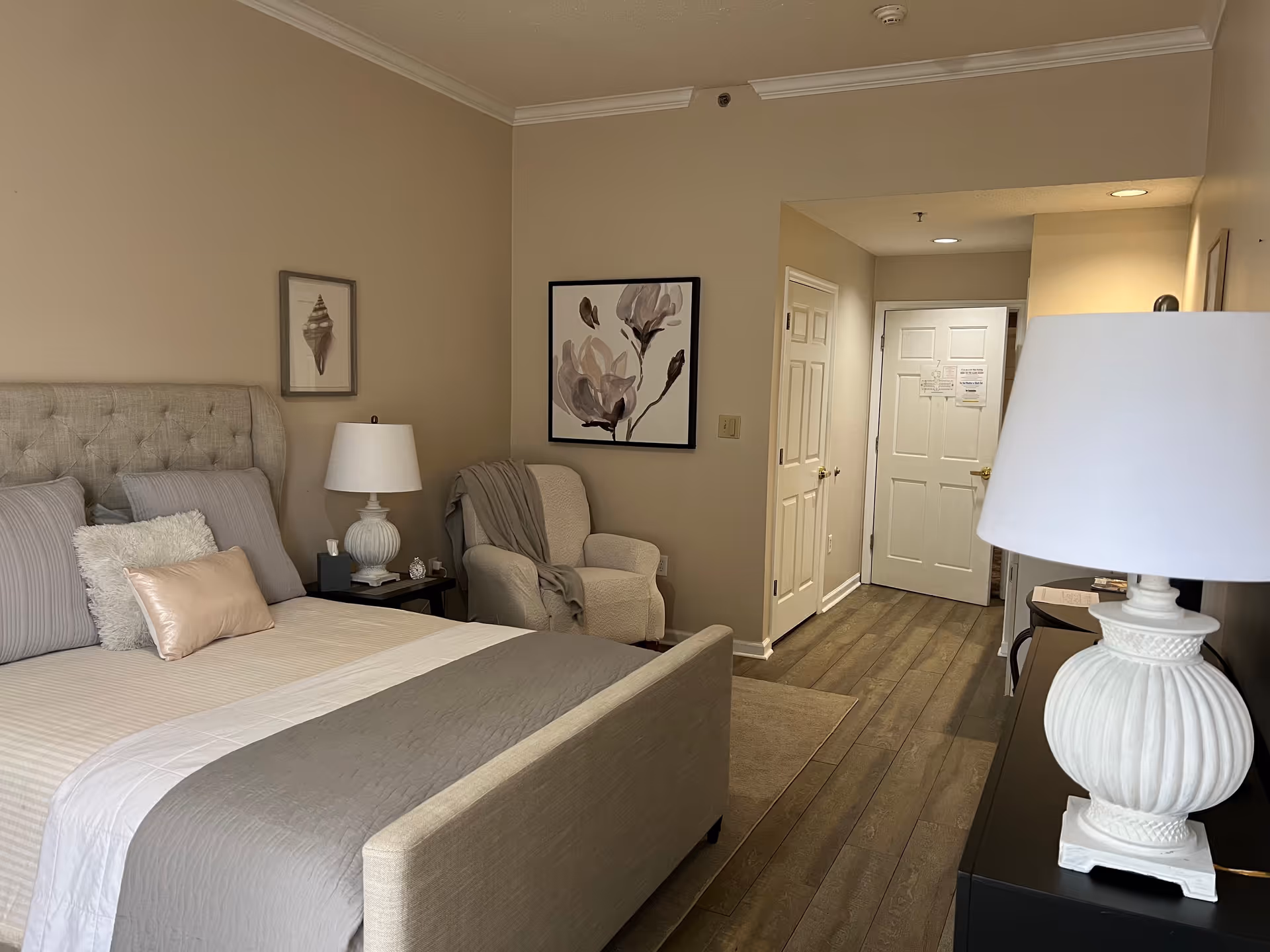 A cozy bedroom in Sumter Retirement Village featuring a neatly made bed with gray and white bedding, a cushioned armchair with a throw blanket, two bedside tables each with a white lamp, framed artwork on the walls, and a wooden floor leading to a closed white door.