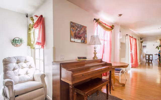 Bright common living area featuring an upright piano, cushioned armchair, round table, lamp, and pink curtains along a hallway.