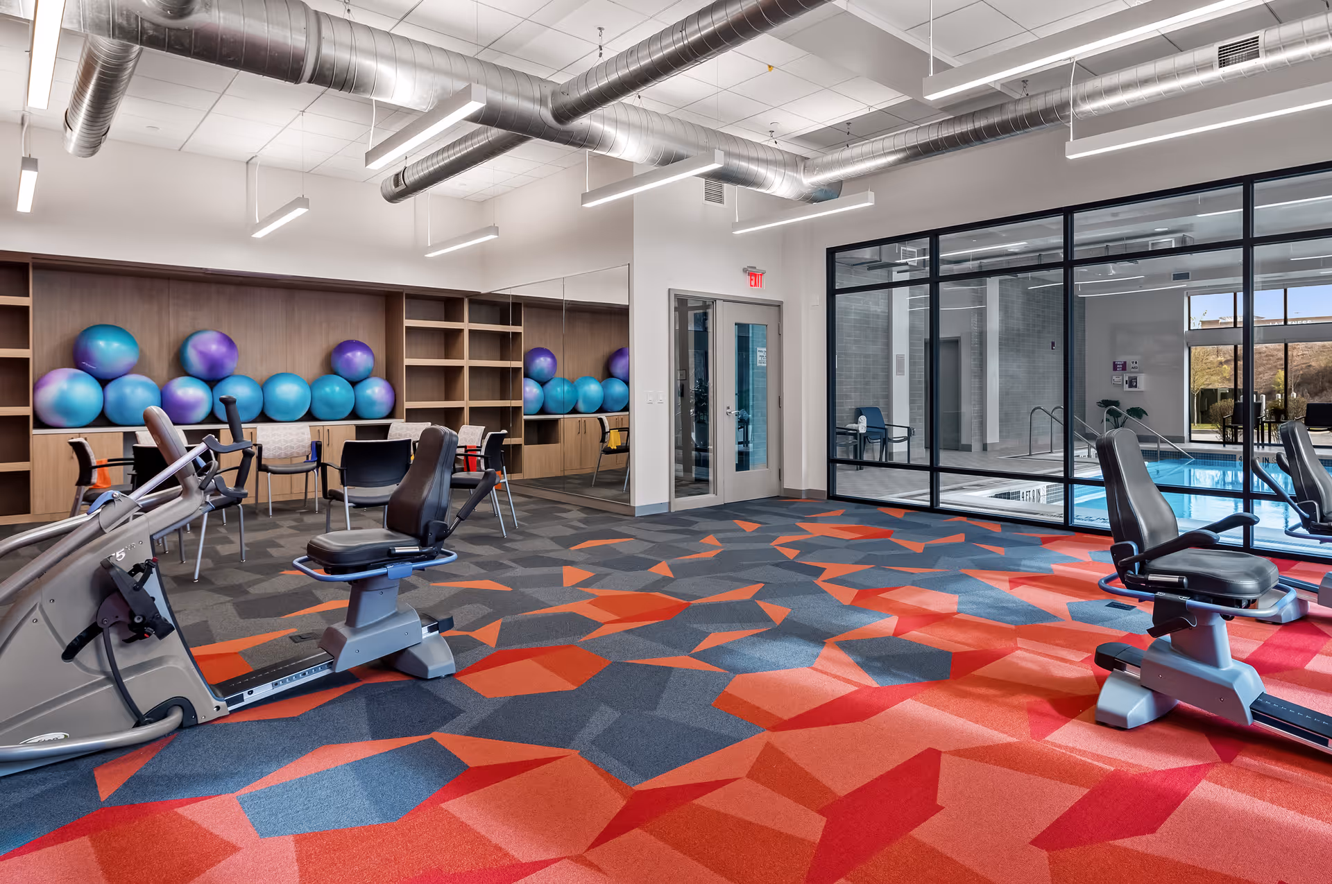 A fitness room with exercise bikes, colorful geometric patterned carpet in shades of red and blue, a wall of shelves holding blue and purple exercise balls, and large windows looking out to an indoor swimming pool area.
