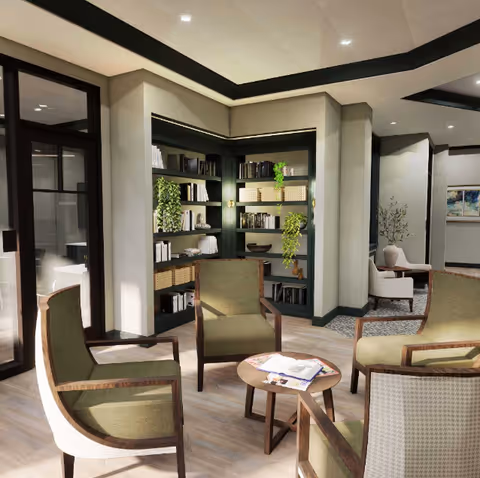 A cozy seating area in a senior living facility with four green cushioned wooden chairs arranged around a small round wooden table with magazines on it. Behind the chairs is a built-in bookshelf filled with books and decorative plants. The room has light-colored walls, wooden flooring, and soft lighting, creating a warm and inviting atmosphere.