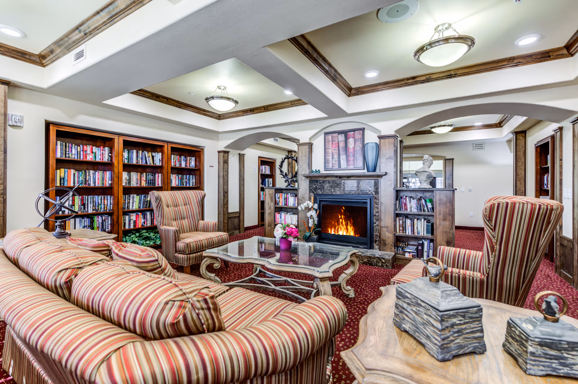 A cozy living room area in Ashton Gardens Gracious Retirement Living featuring striped upholstered sofas and armchairs arranged around a glass coffee table. The room has a lit fireplace with a decorative mantle, bookshelves filled with books, and warm lighting from ceiling fixtures. The carpet is red with a patterned design, and the walls have wood trim and arches.