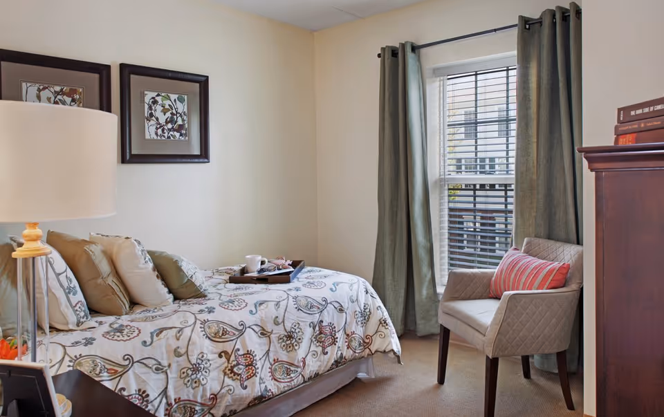 Sunlit bedroom with a patterned bedspread, pillows and tray, bedside lamp, framed artwork, and an upholstered chair by a window with green curtains.