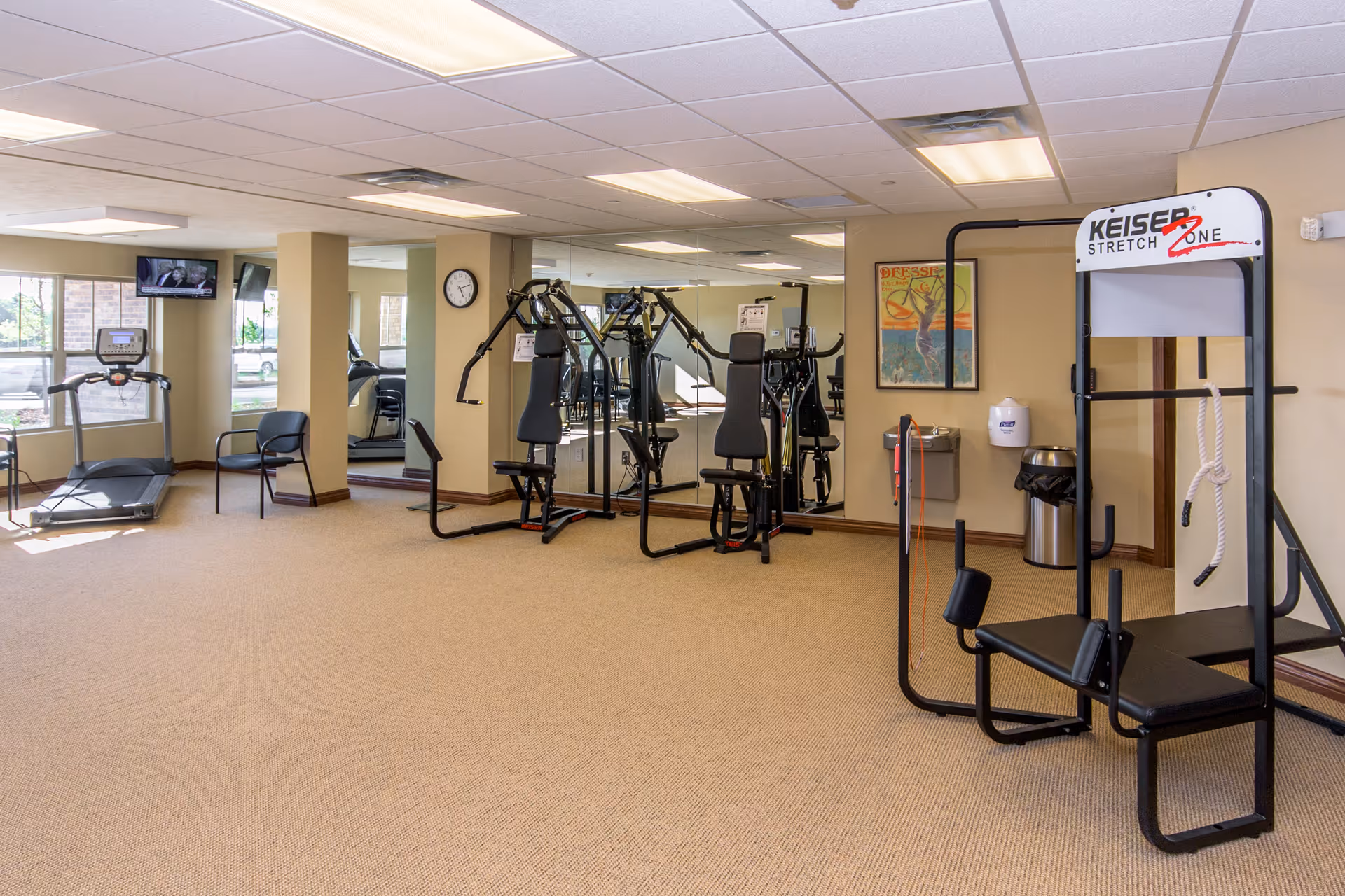 A well-lit fitness room in a senior living facility featuring exercise equipment including a treadmill, weight machines, and a Keiser Stretch Zone machine. The room has beige walls, carpeted flooring, large windows, a wall clock, a water fountain, and a framed poster on the wall.