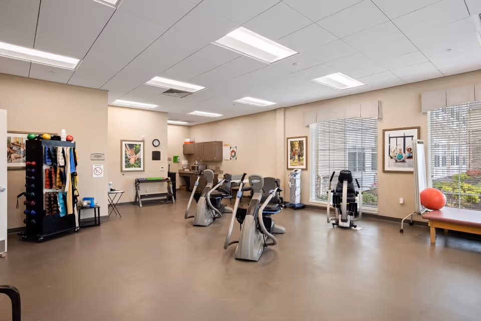 A spacious exercise room in a senior living facility with several stationary exercise bikes, a rack holding various exercise equipment like balls and resistance bands, large windows with blinds letting in natural light, and a bench with a red exercise ball. The walls are beige with framed pictures and there is a small kitchenette area in the back.