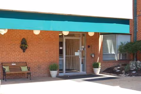 Brick facility entrance with teal canopy, glass doors, bench, and potted plants.