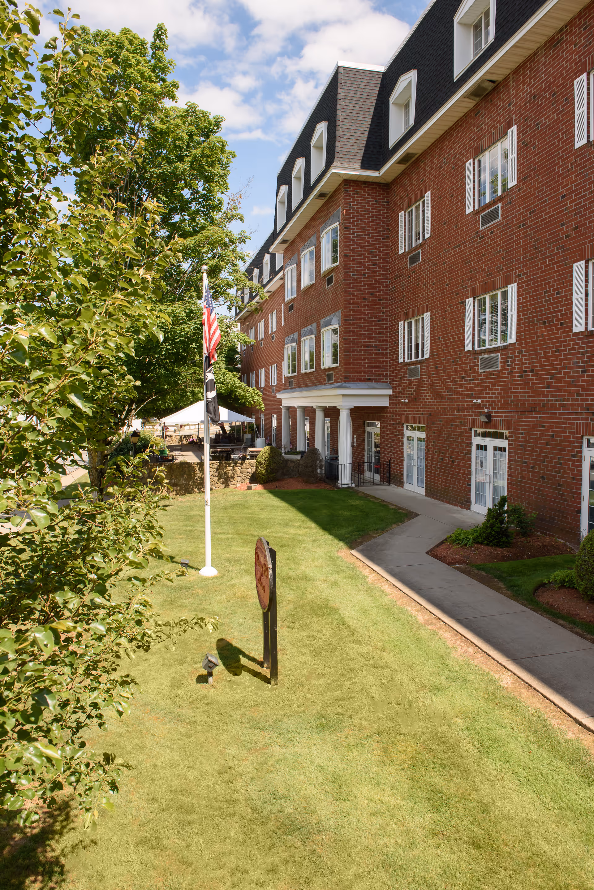 Exterior view of a multi-story red brick building with white-trimmed windows and a black mansard roof. A well-maintained lawn with a flagpole flying the American flag and a POW/MIA flag is in the foreground, along with a sidewalk and some trees. The sky is partly cloudy with blue patches.