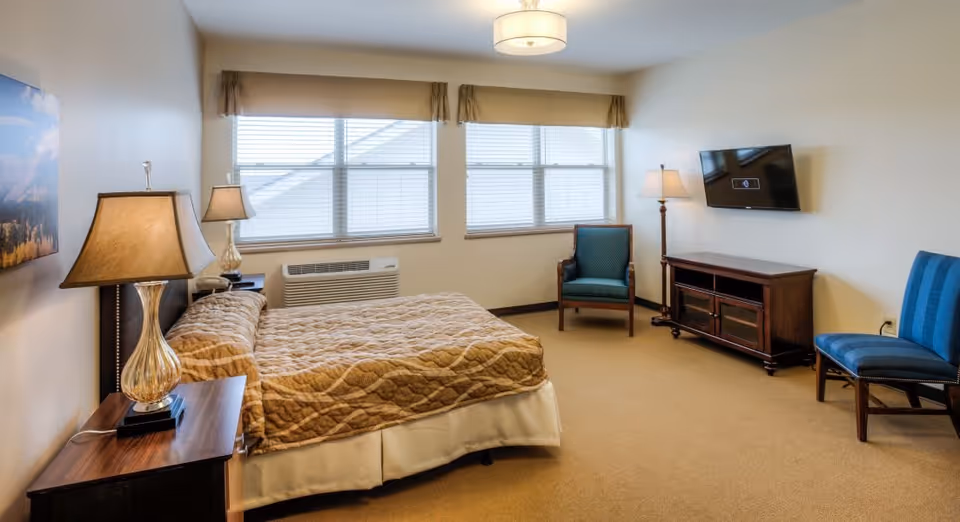 Well-lit senior living bedroom with a bed, bedside lamps, chairs, a wall-mounted TV, and large windows.