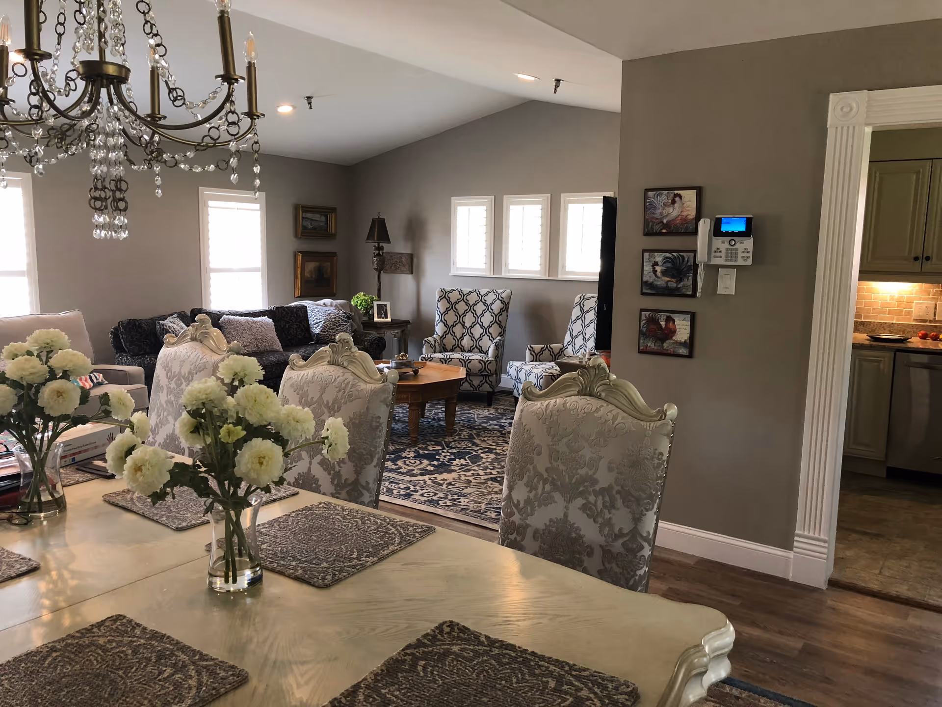 A cozy living and dining area in an assisted living facility. The foreground shows a dining table with ornate chairs and vases of white flowers. In the background, there is a living room with patterned armchairs, a sofa, a wooden coffee table, and decorative wall art. The room has soft lighting and a chandelier hanging above the dining table. A kitchen area is partially visible through a doorway on the right.
