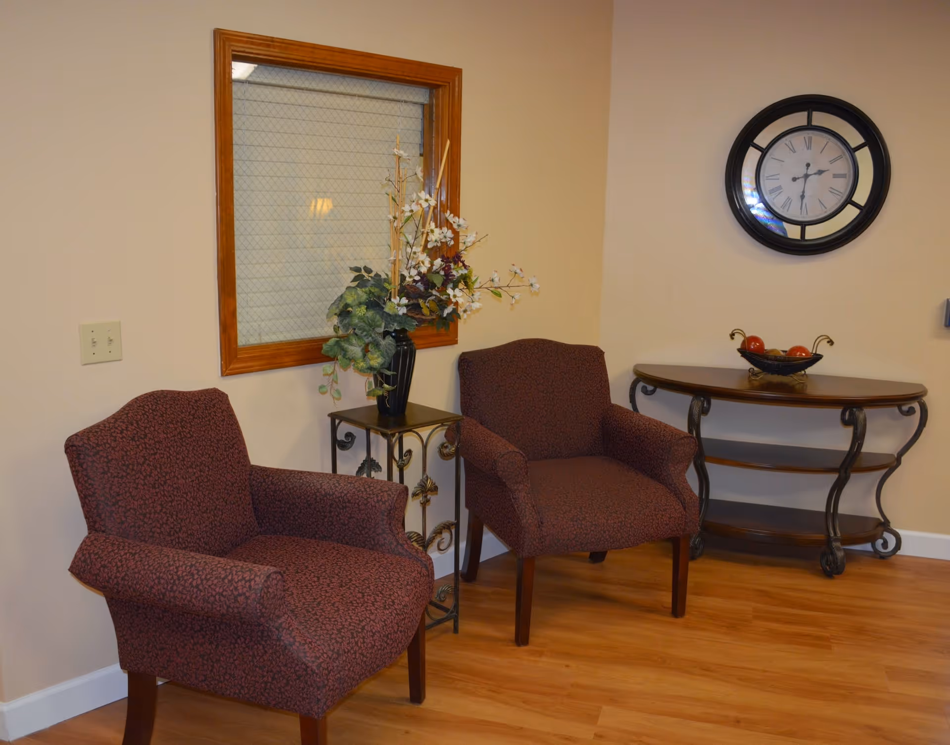 A cozy corner of a room with two patterned armchairs, a small table with a floral arrangement, a wooden console table with a decorative bowl of apples, and a round wall clock above the console table. The floor is wooden and the walls are painted beige.
