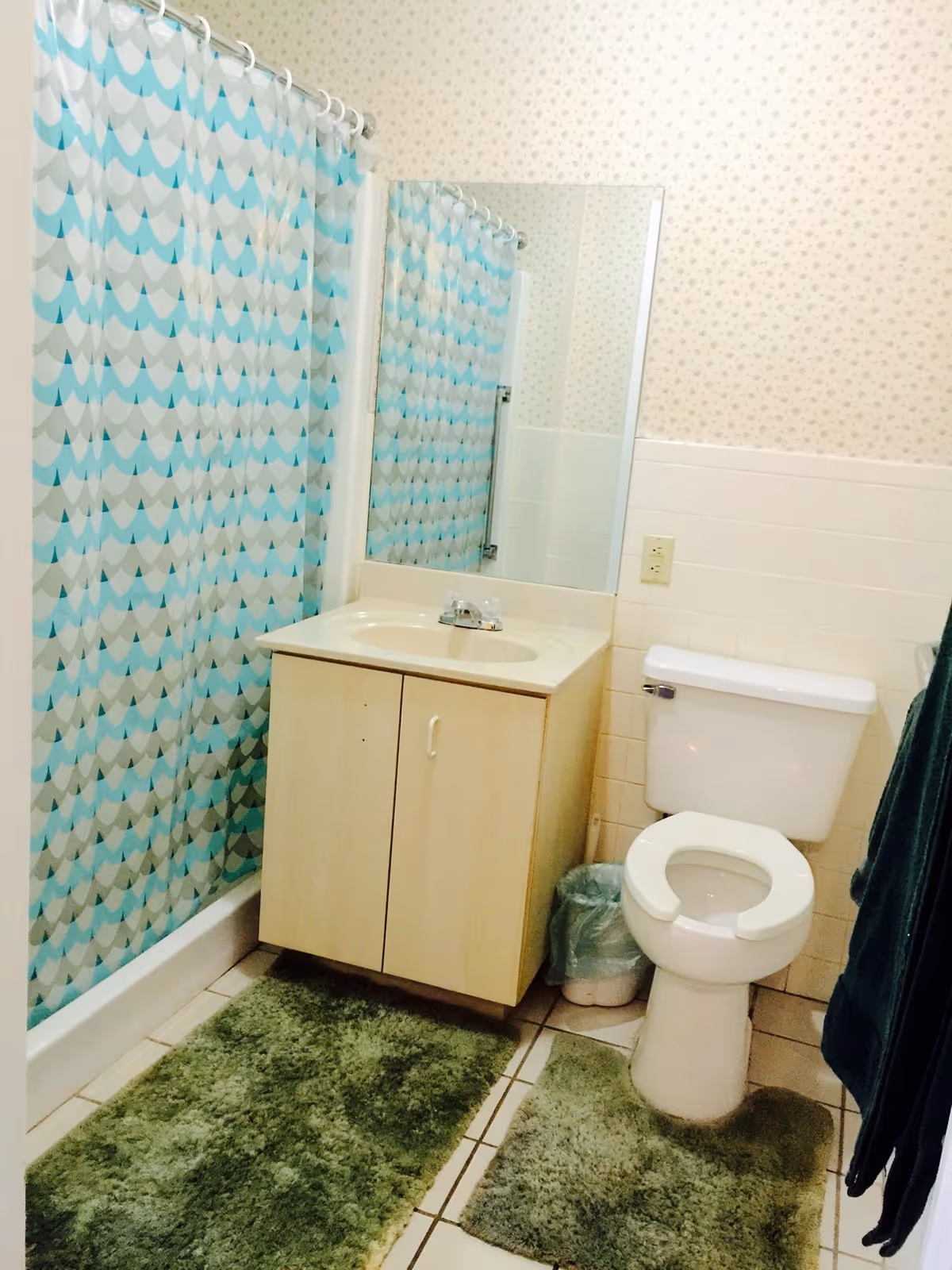 A bathroom with a white toilet, a light wood vanity with a sink and faucet, a large mirror above the sink, a shower with a blue and gray patterned curtain, and green bath rugs on the tiled floor. The walls have a light floral wallpaper and white tile halfway up.