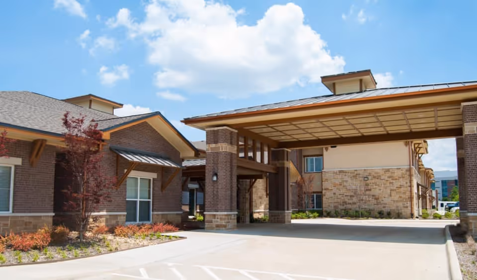 Exterior view of Cedar Bluff Assisted Living & Memory Care facility showing a covered entrance area with brick and stone buildings under a blue sky with some clouds.