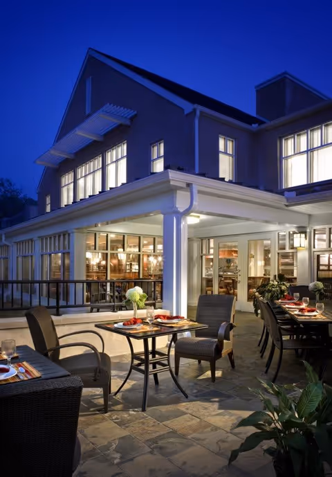 Outdoor patio area of a senior living facility at dusk with tables and chairs set for dining, adjacent to a building with large windows and warm interior lighting.