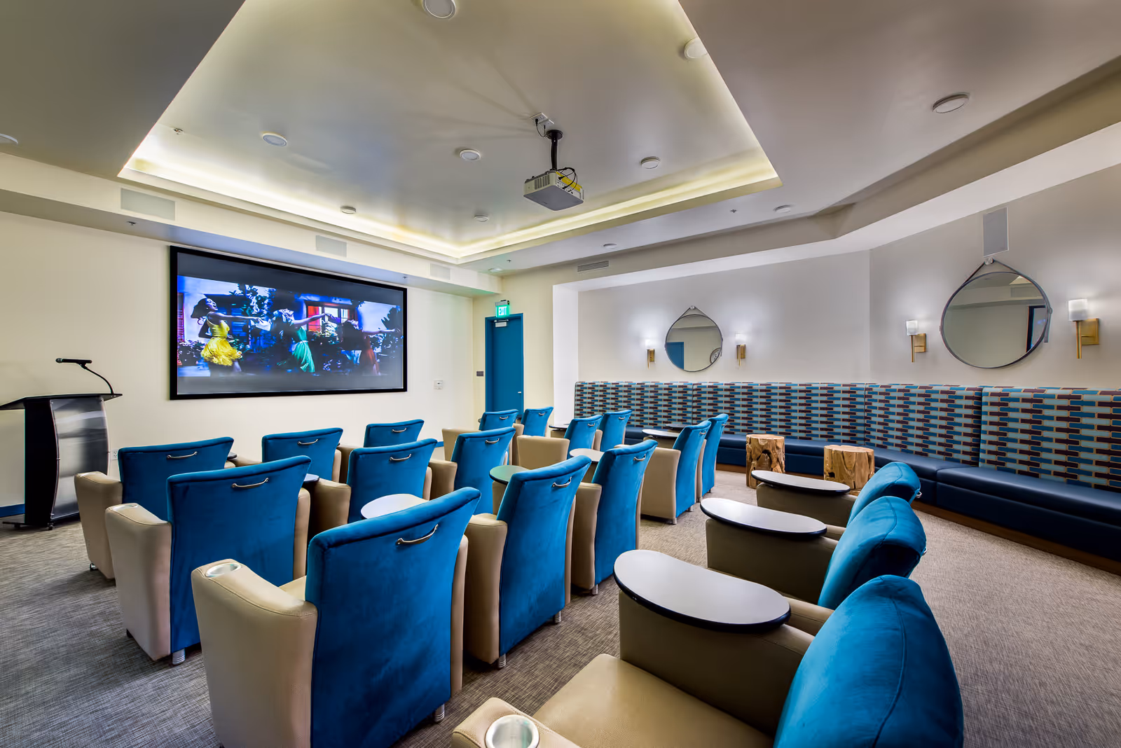 A modern screening room with rows of blue and beige reclining chairs facing a large wall-mounted projection screen and a podium.