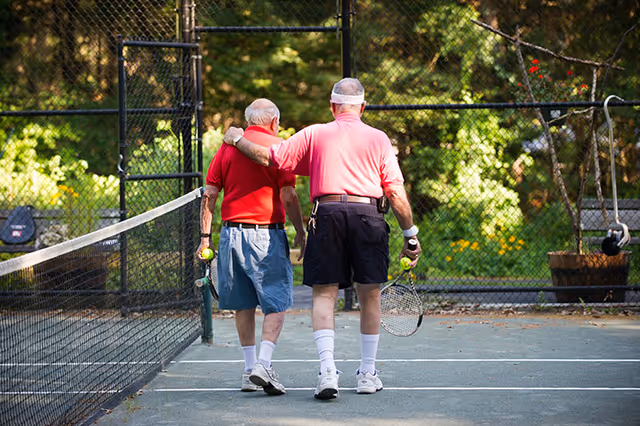 Two elderly men walking side by side on a tennis court, one with his arm around the other's shoulder. Both are holding tennis rackets and tennis balls, dressed in casual sportswear with a green fence and trees in the background.