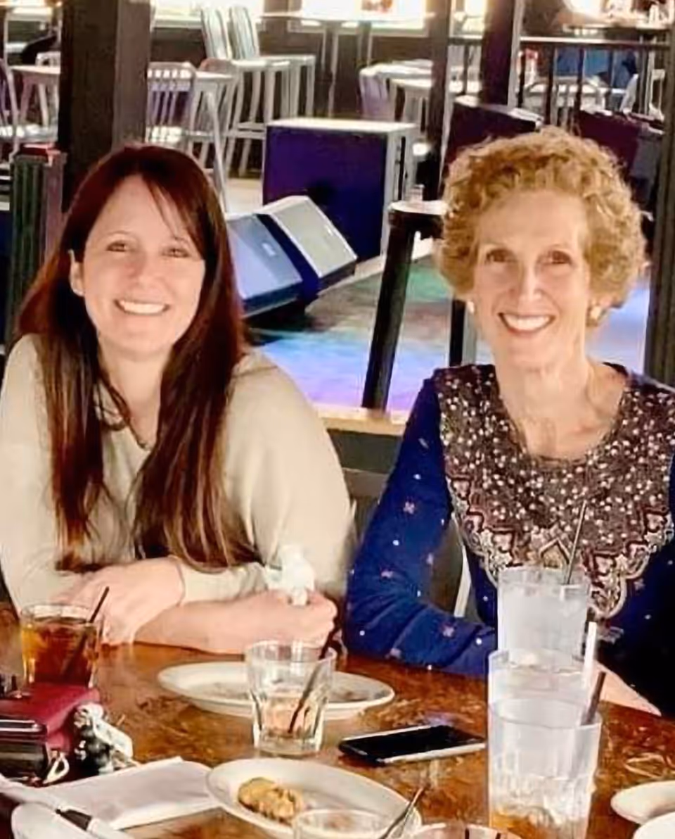 Two women sitting at a wooden table in a dining area, smiling at the camera. The table has several glasses with drinks, a plate with some food, and a smartphone. The background shows chairs and tables in a well-lit room.
