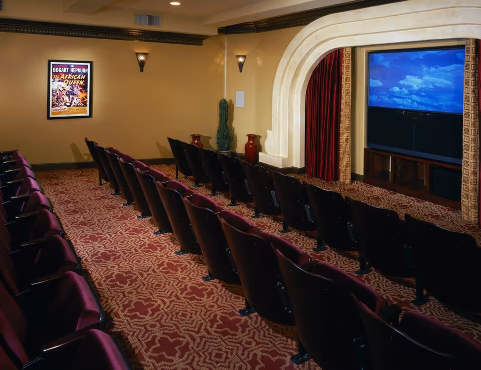 Interior view of a small theater room with rows of maroon cushioned seats facing a large screen displaying a blue sky with clouds. The room has patterned carpet, beige walls, decorative wall sconces, and a framed vintage movie poster for 'The African Queen' featuring Bogart and Hepburn.
