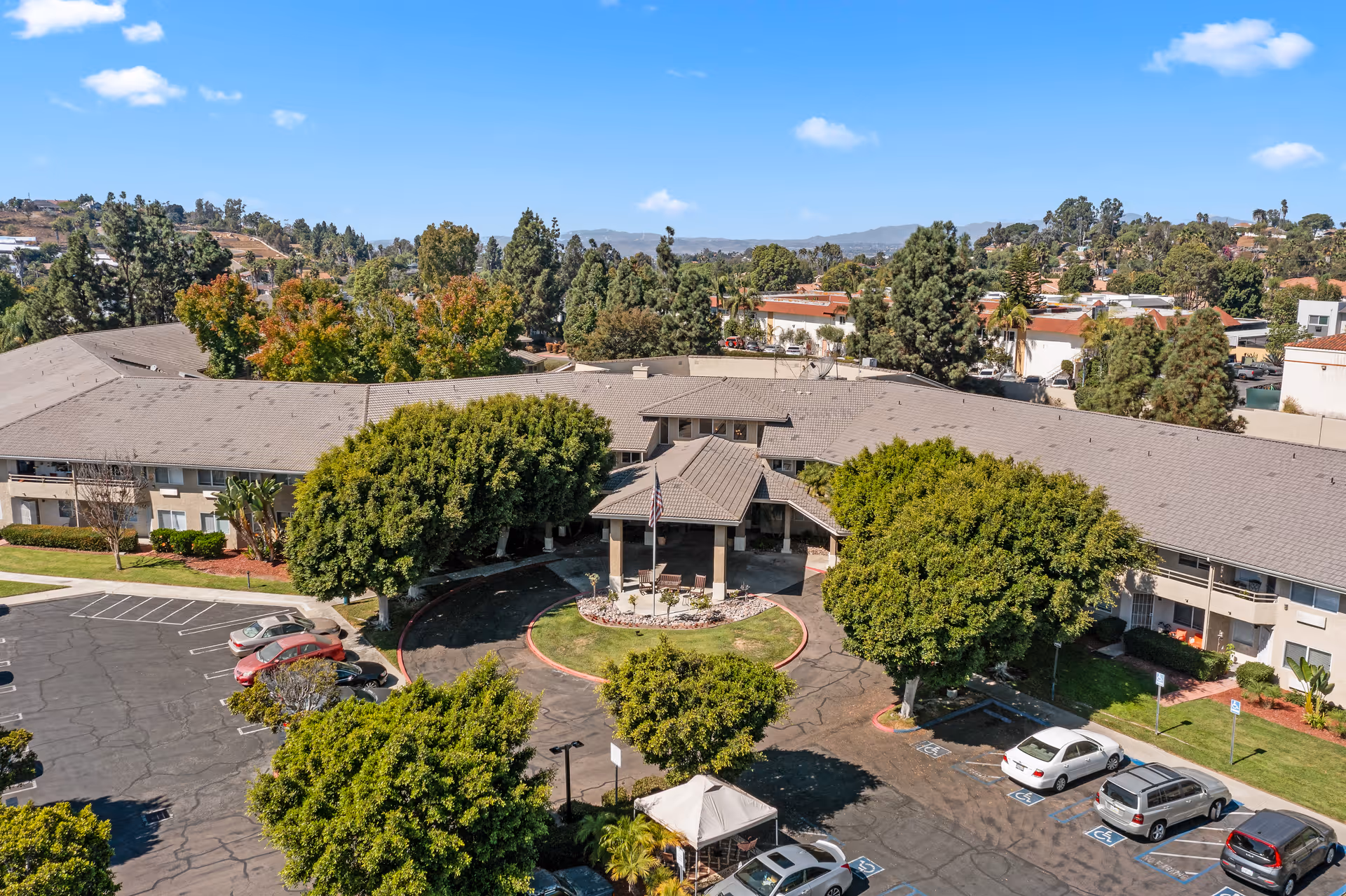 Aerial view of Arcadia Place Senior Living facility showing a large, single-story building with a covered entrance, surrounded by trees and a parking lot with several cars. The sky is clear and blue, and the surrounding area includes more trees and buildings.