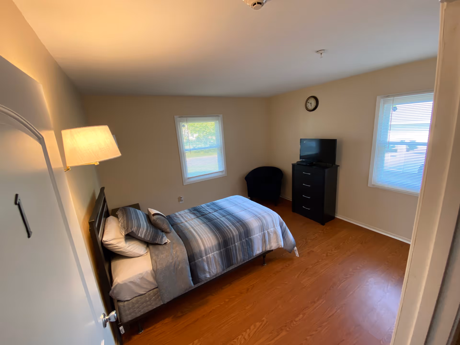 Furnished bedroom with a single bed, wall lamp, dresser with a TV, a chair, and two windows.
