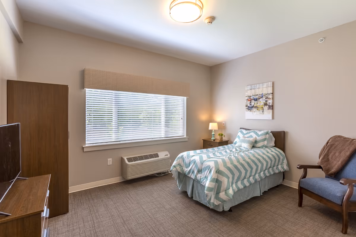 A neatly arranged bedroom in a senior living facility featuring a single bed with a teal and white chevron patterned comforter and pillows. Next to the bed is a wooden nightstand with a small lamp and a plant. A wooden armchair with a brown blanket draped over it is positioned near the bed. Across from the bed is a wooden dresser with a flat-screen TV on top. A large window with blinds and a beige valance allows natural light into the room. The walls are painted light beige, and there is a framed picture of boats hanging above the bed.