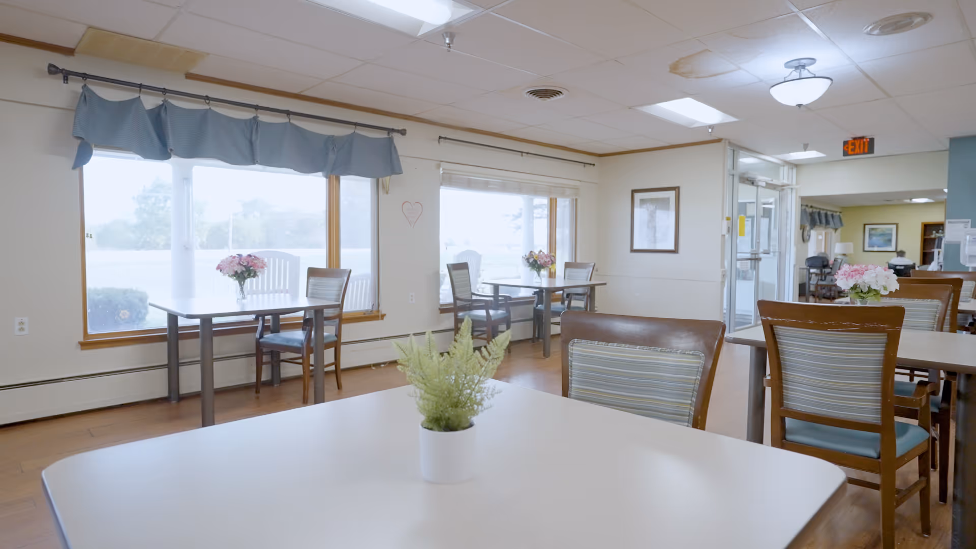 A bright dining area in Oakland Manor with several tables and chairs arranged neatly. Each table has a small floral centerpiece. Large windows with blue valances let in natural light, and the room has light-colored walls and wooden flooring. There is an exit door visible in the background.