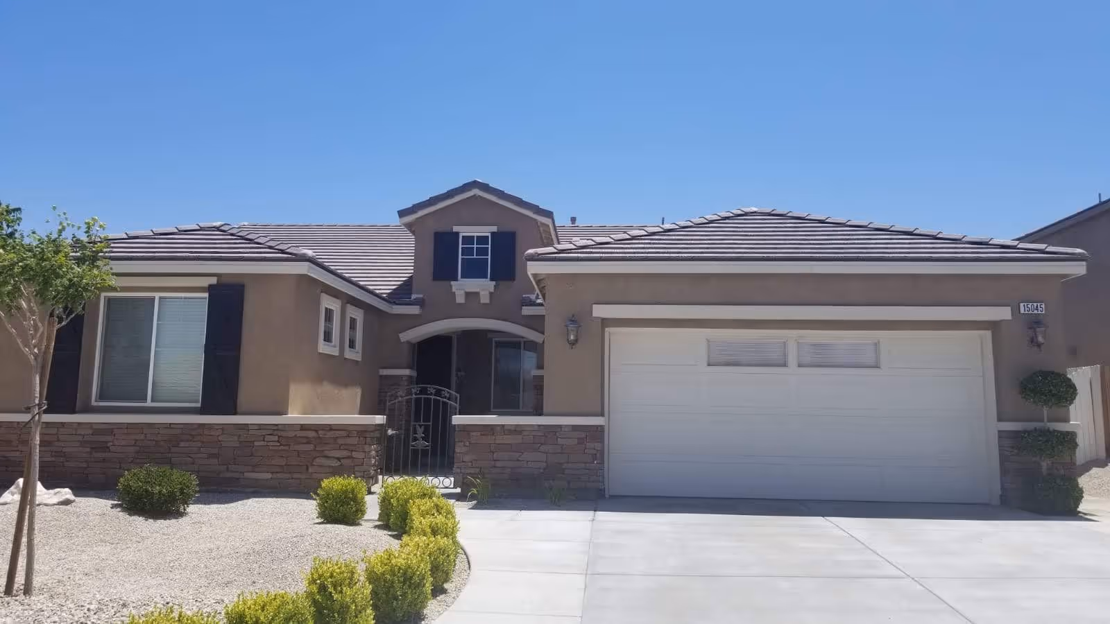 Exterior front view of a single-story house with a two-car garage, beige walls, stone accents, and a small gated front porch. The driveway and front yard have minimal landscaping with small bushes and a tree, under a clear blue sky.