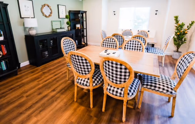 A bright dining room with wooden flooring, featuring a rectangular wooden table surrounded by eight chairs with black and white checkered upholstery. The room has a large window letting in natural light, a black sideboard with decorative items, framed artwork on the walls, and a potted plant in the corner.