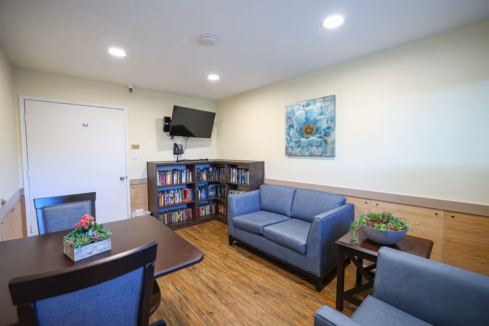 A cozy room with a blue couch, a matching armchair, a wooden table with chairs, a corner bookshelf filled with books, a wall-mounted TV, and a floral painting on the wall. The floor has a wood finish and there are two small plant arrangements on the table and side table.