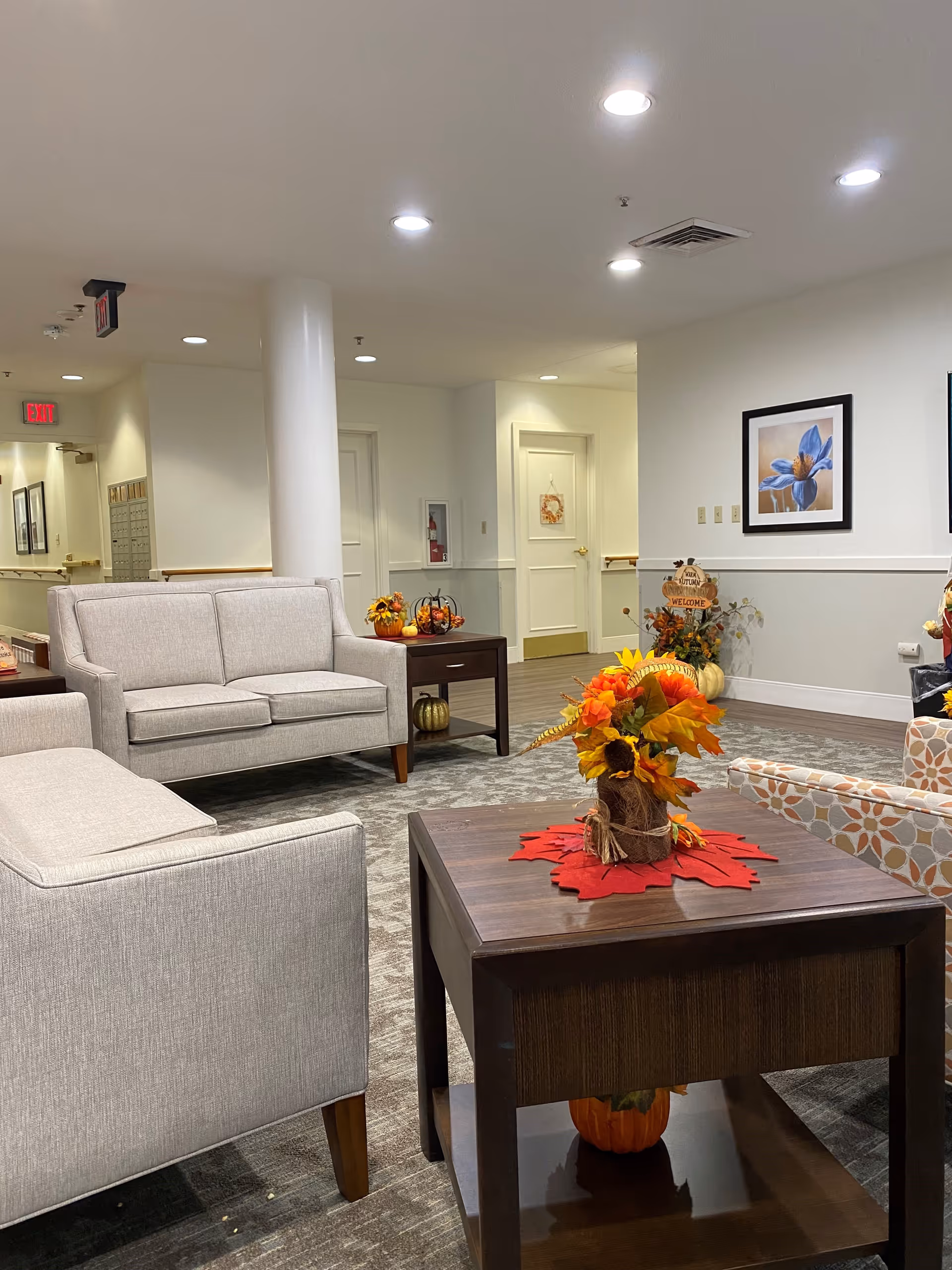 A cozy indoor common area with beige sofas and a patterned armchair arranged around a wooden coffee table decorated with a fall-themed floral centerpiece. The walls are white with framed artwork, and there are autumn decorations including pumpkins and a welcome sign. The space is well-lit with recessed ceiling lights and has a carpeted floor.