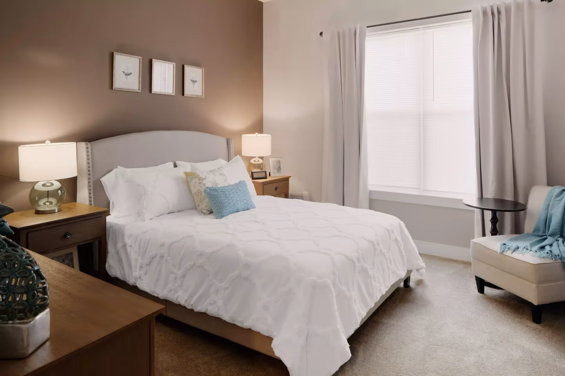 Staged bedroom with a white bed, bedside tables and lamps, a window with curtains, and an upholstered chair.