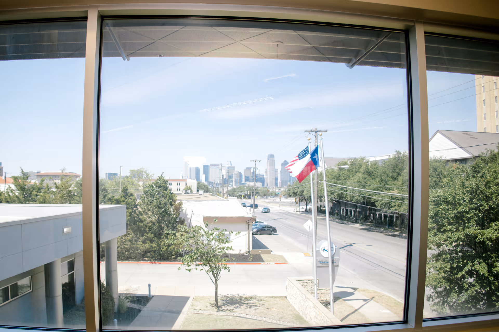View through a large window showing a street with cars, trees, and two flagpoles flying the American and Texas flags. In the distance, a city skyline with tall buildings is visible under a clear blue sky.