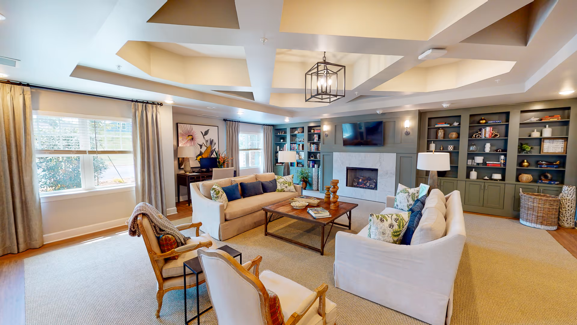 A cozy living room featuring a modern design with comfortable sofas, a wooden coffee table, a fireplace, and bookshelves filled with decorative items. Large windows allow natural light to fill the space, complemented by stylish curtains.