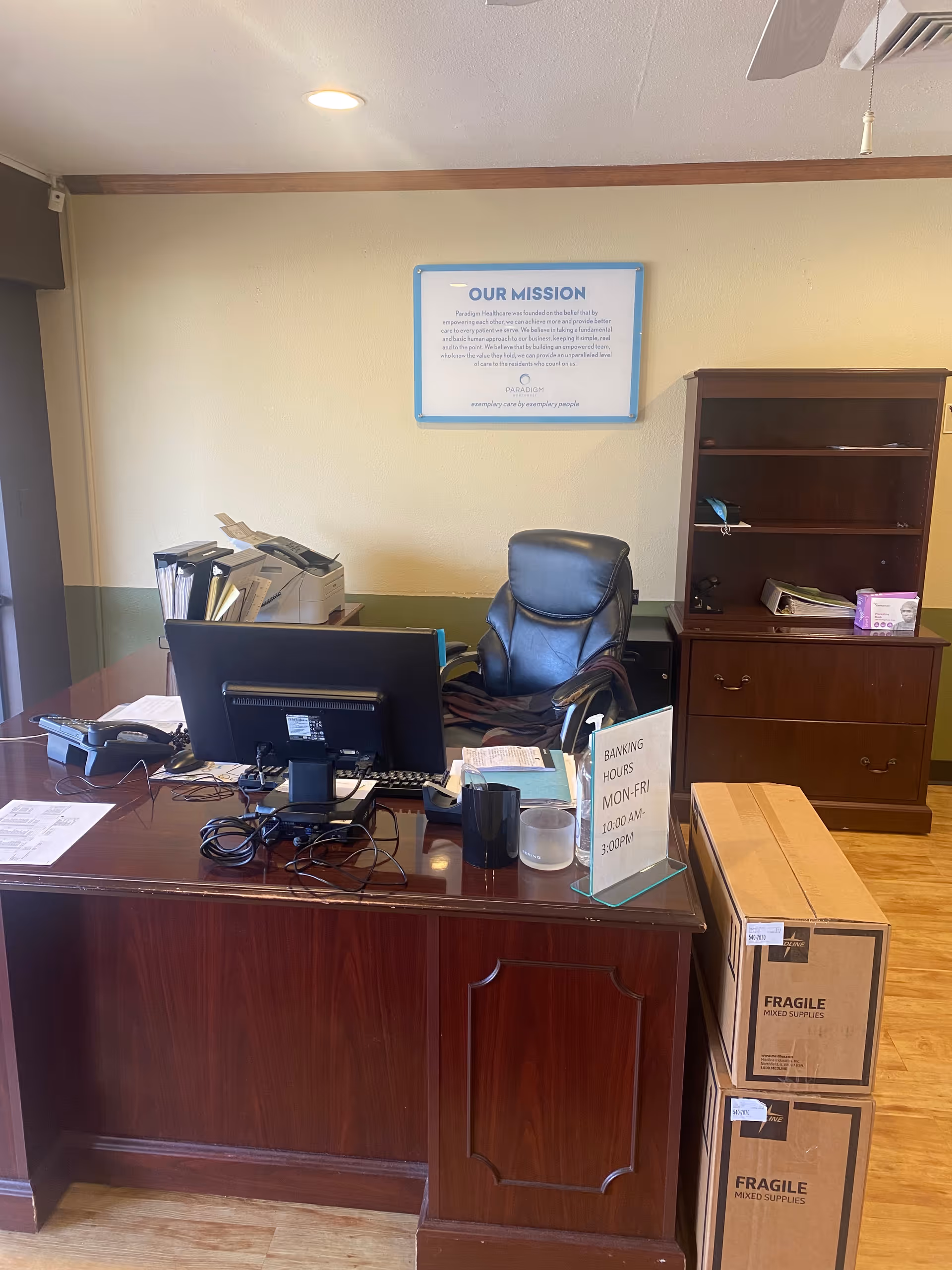 An office space with a wooden desk, a black office chair, a computer monitor, a telephone, and various office supplies. Behind the desk is a wooden bookshelf with some items on it. On the wall above the chair is a framed sign titled 'OUR MISSION'. Two cardboard boxes labeled 'FRAGILE MIXED SUPPLIES' are stacked next to the desk. A small sign on the desk indicates banking hours as Monday to Friday, 10:00 AM to 3:00 PM.