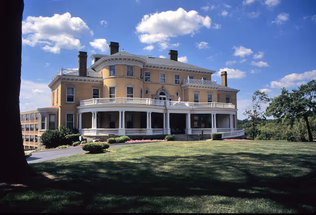 Large historic building with yellow exterior and white wrap-around porch set on a grassy lawn under a blue sky with scattered clouds.