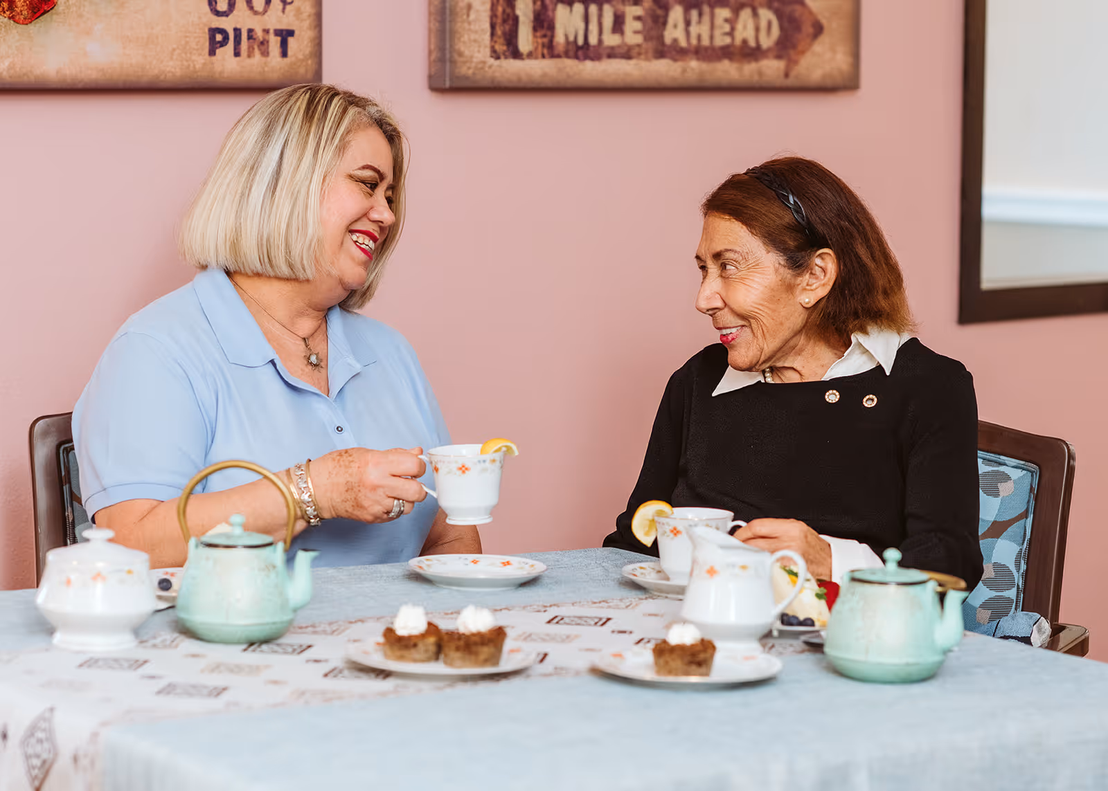Two women sitting at a table enjoying tea together, smiling and engaged in conversation. The table is set with teapots, teacups with lemon slices, plates, and small desserts. The background shows a pink wall with framed artwork.