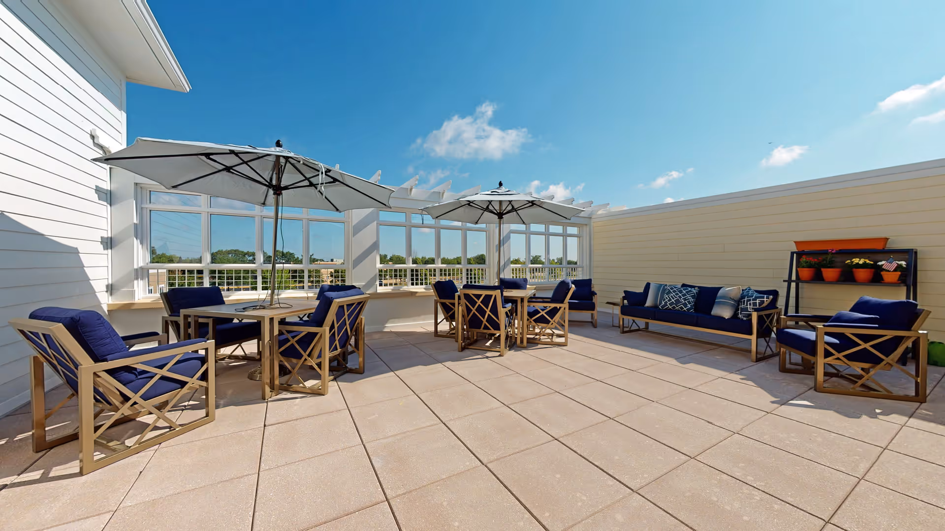 Rooftop outdoor patio with navy-cushioned lounge chairs, tables, umbrellas, and potted plants under a clear blue sky.
